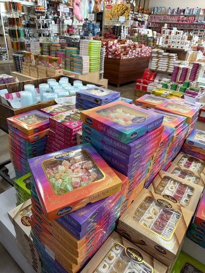 The image depicts a retail store, likely a market or specialty shop, stocked with a variety of food products, primarily Turkish Delight (Lokum) and other confectioneries. The items are displayed in stacks of colorful boxes, some with clear plastic windows revealing the contents. The boxes are branded with "Balkan" and "Türk Lokumu." In the foreground, several stacks of these boxes are prominent, with a particularly large, rainbow-gradient box filled with multi-colored Turkish Delight in the upper left.

In the background, shelves are filled with more packaged goods, including what appear to be more sweets and possibly other food items. To the left, rows of colorful beaded necklaces or similar decorative items are visible hanging from a display. In the middle background, a wooden display crate holds an assortment of cylindrical, pale blue objects, possibly candles. The overall environment suggests a busy retail setting with an emphasis on decorative and edible goods. The image does not contain any people or identifiable landmarks that would specifically place it in the city of Acıpayam, Turkey, or any other specific location.