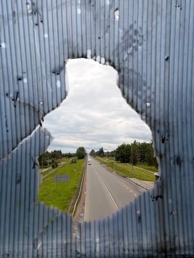 The image captures a view through a jagged hole in a corrugated metal surface. The hole frames a scene of a highway in Maloye Verevo, Russia, under a cloudy sky. The road appears to be a multi-lane highway with vehicles visible in the distance. To the right of the road is a grassy embankment with some trees and streetlights. The corrugated metal in the foreground is blue with dark horizontal lines and shows signs of damage, including some darker smudges and irregular shapes. The lighting suggests it is daytime, possibly late afternoon given the quality of light. The overall impression is one of looking out from a damaged or sheltered structure onto a mundane, everyday scene. There is no discernible text within the image.