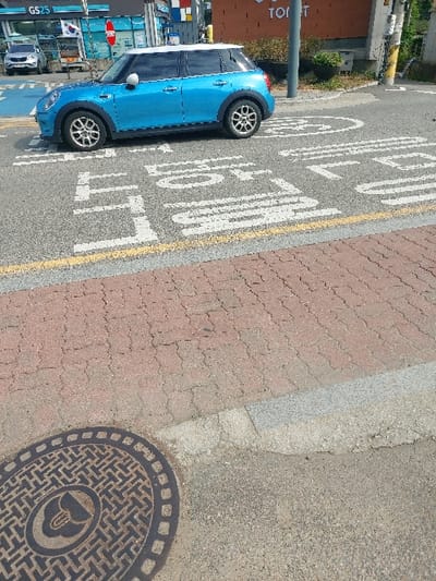 The image depicts a daylight street scene in 강화군, South Korea. A light blue, four-door Mini Cooper with a white roof is stationary on an asphalt road. The vehicle faces left, displaying silver multi-spoke wheels. White Korean road markings on the asphalt ahead of the car read '어린이 보호구역' (Children Protection Zone) and '천천히' (Slowly). A solid yellow line horizontally delineates the road edge. Adjacent to the road is a sidewalk paved with reddish-brown interlocking bricks, separated by a grey concrete curb. In the foreground's lower-left, a circular metal manhole cover with a geometric pattern and a central stylized emblem is visible on a gravel surface. The background features a GS25 convenience store sign, a partially obscured red octagonal stop sign, and a silver SUV parked near the store. A building with a brown facade and a grey utility pole are also visible. No individuals are present in the scene.