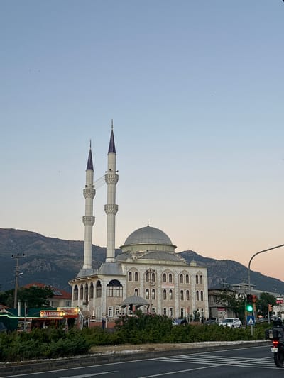 The image depicts a mosque located in Köyceğiz, Turkey. The mosque features a large dome and two minarets with pointed, purple tips. In the background, there are mountains and a street with traffic, including cars and a traffic light that is green. There are also businesses visible, with some written in Turkish, such as "ÇIĞLA KOKOREÇ". The sky is a gradient of light blue to light orange, suggesting either dawn or dusk. The camera angle is slightly tilted.