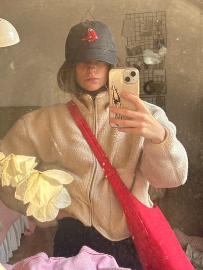 A person takes a mirror selfie, wearing a gray baseball cap with a red logo, a cream-colored fleece jacket, and a red crossbody bag. They hold an iPhone with a sticker on the back. Part of a grid wall organizer and some other items are visible in the background. A plant with large white flowers is in the foreground. The overall lighting appears soft and warm. The mirror's surface is somewhat dirty or textured.
