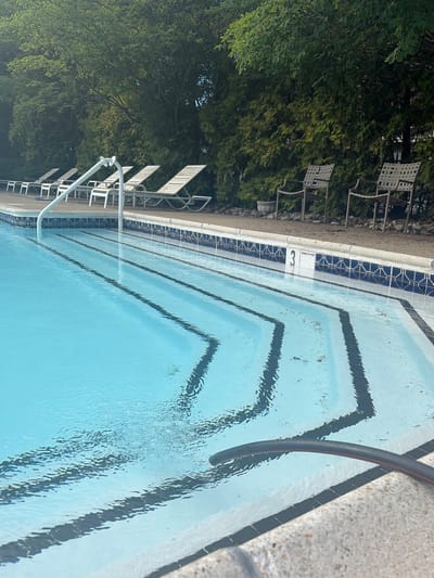The image depicts a swimming pool with a ladder and a hose extending into the water. The pool has black lane markers and is surrounded by a tiled border and concrete coping. Lounge chairs are visible alongside the pool, with trees in the background. The location is indicated as Edison, United States of America, but there are no further specific location details visible in the image. There are no people visible in the image.