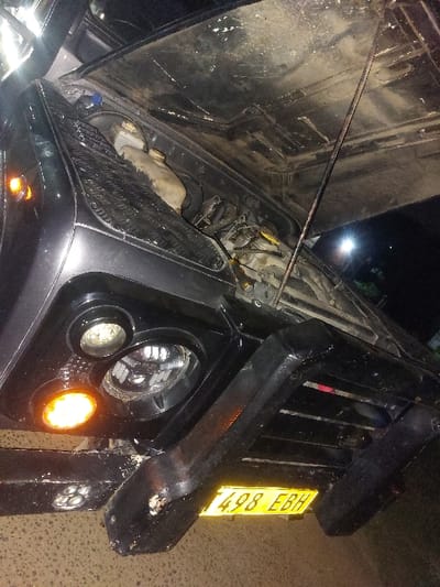 The scene, located in Msata, Tanzania, depicts the front left section of a dark-colored utility vehicle, consistent with a Land Rover Defender, at night. The vehicle's hood is open and supported by a metal rod, revealing a soiled engine bay. Visible engine components include a light-colored, soiled coolant reservoir and a washer fluid reservoir with a blue cap. A yellow-handled tool, resembling a screwdriver, rests amidst the engine's wiring and hoses. The front left headlight assembly features a main circular headlight, a smaller auxiliary light, and an illuminated amber turn signal. A robust, dark-colored front bumper, showing signs of paint chipping and wear, is present. A yellow license plate, T498 EBH, is affixed below the bumper, indicating Tanzanian registration. The vehicle rests on an asphalt or paved surface, with a distant bright light source visible against the dark background. No individuals are present.
