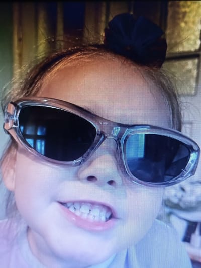 The image shows a close-up of a young girl wearing oversized, clear-framed sunglasses and a dark blue hair accessory. Her mouth is open, revealing her teeth, and she appears to be looking forward. The background is blurred, but it appears to be an indoor setting with vertical wooden elements and a window. There are no discernible elements in the image that would allow for the identification of the specific location within the city of Yampil, Ukraine.