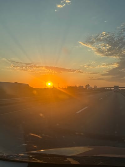 The image depicts a sunset viewed from the perspective of a vehicle traveling on a highway in Madera, California. The sun is low on the horizon, casting a warm orange glow across the sky and illuminating the clouds with golden hues. Rays of sunlight are visible emanating from the sun, creating a dramatic effect. The highway stretches into the distance, with other vehicles, including trucks and cars, visible on the road. The landscape to the left of the highway appears somewhat arid, with low vegetation and some structures. The foreground shows the interior of the vehicle, likely the dashboard and windshield, with reflections from the sunset. The overall impression is one of a scenic drive at dusk on a clear day.