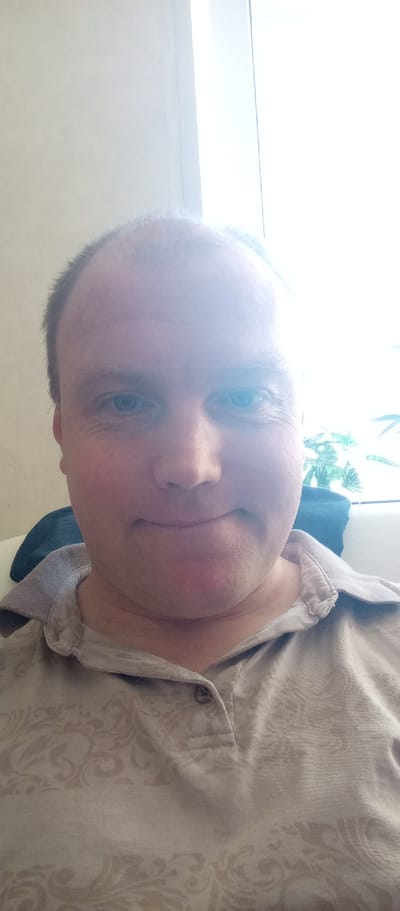 The image is a close-up selfie of a man in Стулово, Russia. He is a bald, middle-aged man with blue eyes and fair skin. He is wearing a light brown collared shirt with a subtle patterned design. The background is mostly a brightly lit window, suggesting it is daytime. A hint of green foliage is visible outside the window, possibly indicating plants or trees. The man is looking directly at the camera with a slight, closed-mouth smile, conveying a neutral or pleasant emotion. The lighting is harsh, creating some overexposure and glare on his forehead and the upper part of the image, possibly from direct sunlight coming through the window. The overall scene is domestic, likely taken indoors in a living space. There is no visible text in the image.
