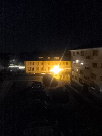 The image depicts a nighttime scene in Lauterbourg, France. In the background, two apartment buildings are visible. The building on the left is illuminated by a bright street lamp and the interior lights of its windows. The building on the right, a taller concrete structure, also has several illuminated windows. In the foreground, a parking area is partially visible, with several cars parked in the darkness. A brightly lit object, likely a streetlight, casts long shadows across the scene. The overall impression is one of a quiet, residential area at night.