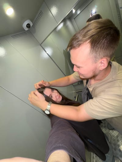 In what appears to be an elevator, a young man with light brown hair and a short beard, wearing a tan t-shirt, is seen interacting with a young woman. He has his hands in her dark brown hair, pulling it slightly. The woman, with long dark hair, is looking towards the camera with a slight smile. She is wearing a dark long-sleeved shirt and a silver watch on her left wrist. The elevator walls are a neutral gray color with horizontal lines. A security camera is visible on the ceiling in the upper left corner of the image. The floor of the elevator is a metal grate. The overall impression is of a playful or casual interaction. The location is Shul'gino, Russia.