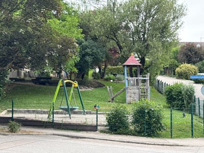 This media file depicts a children's playground nestled within a leafy, park-like setting. The playground features a swing set with lime green A-frame supports and a blue tire swing. Adjacent to the swing set is a wooden climbing structure with a red-capped roof, a slide, and a climbing wall with colorful handholds. A sandpit, enclosed by a low stone wall and a green metal fence, is also visible. The area is covered in well-maintained green grass, with mature trees providing ample shade. In the background, a road or pathway winds uphill, bordered by a dark green metal fence and lush foliage. A signpost with the text "Etzmattenstraße" is partially visible on the right side of the image, indicating the location as Etzmattenstraße. The scene appears to be set during the daytime, under an overcast sky, suggesting a mild or slightly cloudy day. The playground is empty, implying a quiet moment, perhaps before children arrive or after they have left. The overall impression is one of a peaceful, well-equipped outdoor space designed for children's recreation.
