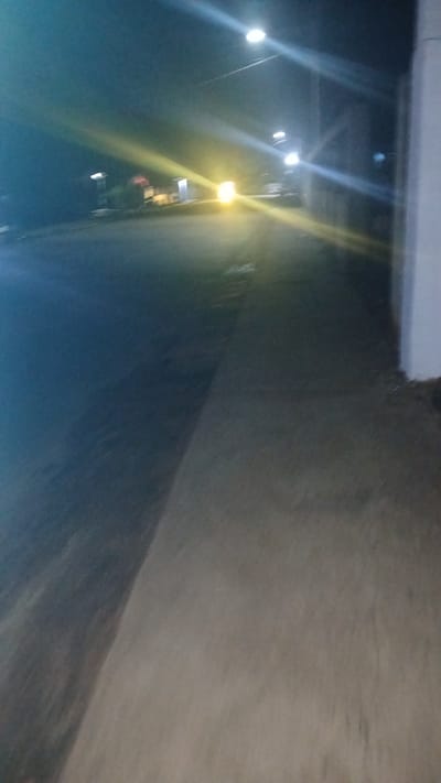 The image shows an outdoor scene at night in what appears to be El Samán, Venezuela. The perspective is angled, suggesting the photo may have been taken handheld and tilted. Streetlights illuminate a road or pathway running along a concrete structure, possibly a wall or building. The lighting is predominantly dim, with the bright streetlights creating intense lens flares. No people or other identifiable figures are visible. The overall environment suggests an urban or suburban setting at night.