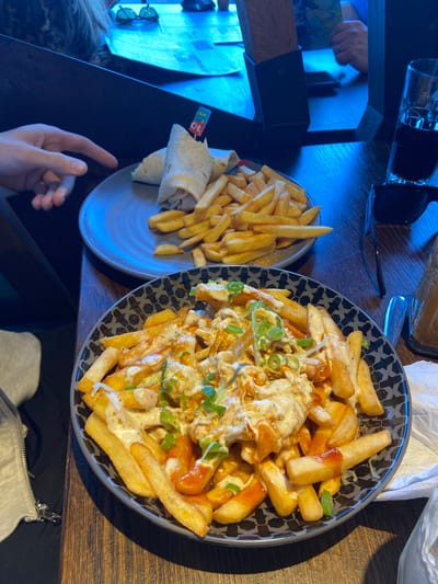 The image shows a table in what appears to be a restaurant setting. On the table are two plates of food: one containing french fries with a wrap, and the other containing french fries topped with sauce, cheese, and green onions. There are glasses and other objects on the table, as well as a hand reaching into the frame. The location is Elmbridge, United Kingdom, based on the geotag.