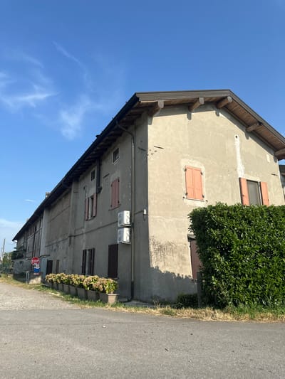 The image shows the side of a weathered, multi-story building in what appears to be a rural setting. The building's facade is made of concrete and shows signs of age, with some discoloration and repairs. Several windows are visible, some with closed wooden shutters painted a reddish-brown. Above some windows, air conditioning units are mounted. The roof has exposed wooden beams and is covered with grey tiles. Adjacent to the building is a very dense, dark green hedge. The scene is bordered by an asphalt road on the right, with dry grass and some plants in planters along the building's base. A stop sign is visible on a pole near the road, indicating a street intersection. The sky is a clear, vibrant blue with a few wispy clouds. The lighting suggests it is daytime, likely mid-morning or afternoon, with the sun casting shadows from the hedge onto the building. The overall impression is one of a lived-in, possibly older structure in a quiet, perhaps Italian, location. The image is taken from a low angle, looking upwards towards the building and sky.