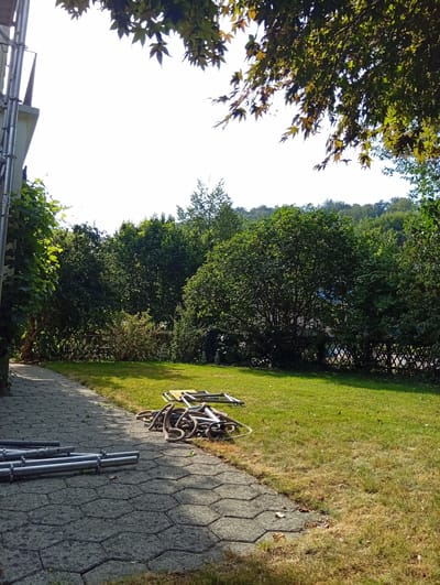 The image depicts a sunny outdoor scene, likely in a garden or park area, possibly in Zofingen, Switzerland. A paved pathway, made of interlocking hexagonal grey stones, runs along the left side of the frame and curves into the distance. To the right of the pathway is a well-maintained green lawn. On the pathway, several metallic cylindrical objects are neatly arranged, appearing to be part of a construction or scaffolding structure. Further into the lawn, a tangled pile of thick, dark ropes is visible, along with some metal scaffolding components. The background is dominated by dense, lush green trees and foliage, with a hint of a hill or slope covered in trees at the far end. The sky is bright and clear, suggesting a pleasant day. Overhanging branches with green and some reddish-brown leaves frame the top of the image. No people are present in the scene.