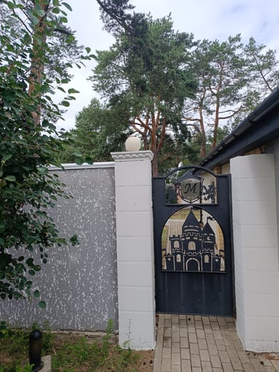 The image shows a black metal gate with a castle silhouette and a heraldic crest with the letter "M" on it. The gate is flanked by white pillars and part of a gray, textured wall on the left, and a white building with a dark gutter on the right. A paved walkway leads up to the gate. Green trees, including pine trees, are visible in the background, with a cloudy sky overhead. A bush with dark berries is in the foreground on the left. The scene is located in Старобінскі сельскі Савет, Belarus.