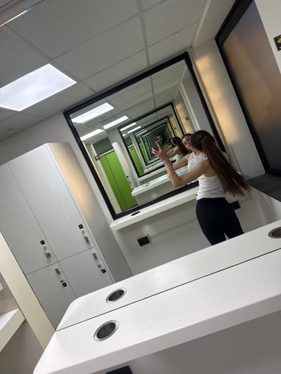 The image depicts a female individual engaged in a mirror selfie within a modern changing room or locker facility located in Lucca, Italy. The subject, a woman with long brown hair pulled into a ponytail, wears a white sleeveless top and dark form-fitting trousers. She is captured holding a smartphone, identifiable as an iPhone with multiple rear cameras, in her raised right hand, angled to photograph her own reflection.

The primary visual element is a large, black-framed mirror positioned centrally above a white counter. This mirror reflects an opposing wall that also contains mirrors, generating an "infinity mirror" effect where successive reflections recede into the distance. The white counter features two integrated circular metallic-rimmed holders. To the left of the frame, a bank of white or light wood-toned lockers equipped with silver-colored locks and dark square labels is visible. The ceiling is a white drop-tile grid system with multiple bright, rectangular fluorescent light panels. Within the depth of the mirror reflections, bright green doors or partitions, indicative of individual changing or shower stalls, are discernible. The overall environment is brightly illuminated by artificial lighting.