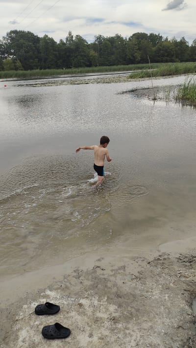 In the city of Ramon, Russia, on a cloudy day, a young boy is walking in the shallow water of a lake. He is wearing black swim trunks with a blue stripe. The boy is facing away from the camera, and his left arm is extended to his side. His movement is creating ripples in the water. The lake is surrounded by a dense forest of green trees and reeds. In the foreground, two black Croc-style shoes are resting on the sandy shore. The sky is overcast with some clouds visible.