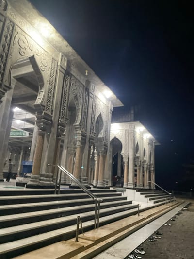 An ornately designed, possibly religious, structure is captured at night. The architecture features arched openings, decorative moldings, and detailed pillars. A staircase with metal railings runs along the side, parallel to the structure. The building appears to be constructed of a light-colored material, possibly stone or concrete. The background is a dark night sky.