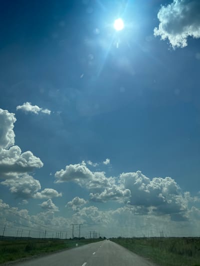 The image captures a bright, sunny day with a clear blue sky filled with scattered white cumulus clouds. The sun is high and radiant, creating lens flare effects and bright highlights in the upper portion of the image. Below the sky is a flat, open landscape, likely a steppe, with a paved road extending into the distance. The road is marked with white lines and appears to be a highway. Along the sides of the road, there is green grass and vegetation. In the middle ground, a series of tall electricity pylons with wires strung between them stretches across the horizon, indicating infrastructure in a rural or semi-rural area. The scene appears to be a view from a moving vehicle looking forward along the road, with the sky occupying the majority of the frame. The location is identified as Ульянов ауылдық округі, Kazakhstan.