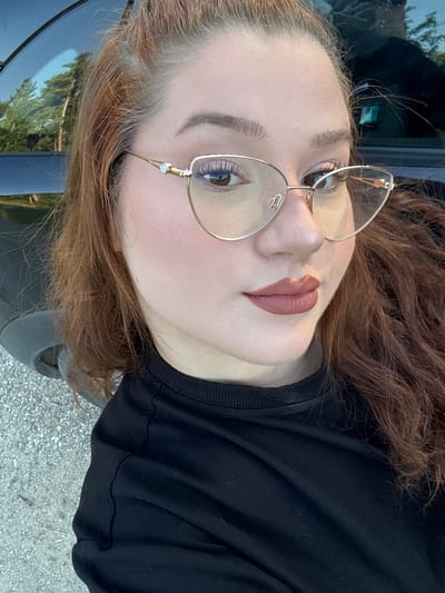 A selfie of a woman with red hair and glasses. She is wearing a black shirt. The background appears to be outdoors, possibly near a car. It is difficult to determine if the location is Malko Kirilovo, Bulgaria without additional context or landmarks.