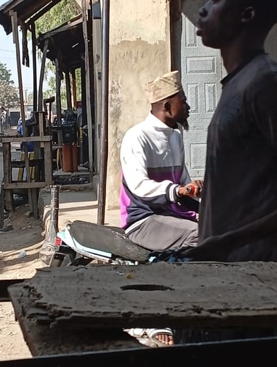 The scene is set in Kofar Ruwa, Nigeria, depicting an outdoor street or market environment under bright daylight. In the immediate foreground, a dark, rough, rectangular surface, possibly concrete or wood, partially obscures the lower portion of the image. Beneath this surface, a gap reveals a person's bare feet clad in white and black striped sandals.
In the midground, an adult male is seated on the black, textured seat of a motorcycle or scooter, oriented towards the right. He wears a white long-sleeved top with a horizontal purple or dark magenta stripe across the chest and dark lower garments. A light-colored, patterned cylindrical cap covers his head, and he has a short, dark beard. His right hand is visible, holding a small red object. To his right, an obscured adult male stands in the foreground, largely in silhouette, wearing a dark shirt. Only his upper torso and the left profile of his head are visible, with his mouth slightly open.
Behind the seated man, a light-colored, weathered building wall is visible, featuring a patterned, recessed metal door further to the right. To the left of the seated man, a rustic wooden structure with a flat roof, possibly a stall or shelter, stands. Visible within and around this structure are several wooden chairs and other indeterminate objects. The background to the left shows a dusty street or open area with faint outlines of distant buildings, trees, and a person dressed in blue.