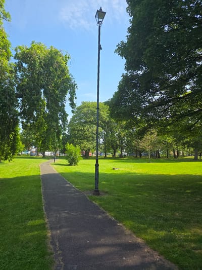 The image shows a park with a paved pathway winding through a lush green lawn. Tall trees with dense foliage line the perimeter of the park, casting shadows on the grass. A vintage-style streetlamp stands prominently beside the pathway, its black metal post and lantern-shaped lamp adding a touch of old-world charm. The sky above is a vibrant blue, dotted with scattered white clouds. In the distance, buildings can be seen peeking through the trees, suggesting an urban setting. This park is likely in the city of Stockton-on-Tees, United Kingdom, given its typical English park characteristics. There are no people visible in the image, and the scene appears to be one of peaceful tranquility.