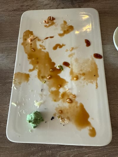 The image shows a rectangular white plate, likely ceramic, containing remnants of food. There are visible traces of a dark brown sauce, possibly soy sauce or teriyaki sauce, along with scattered small food particles. A ball of green wasabi is located in the lower right corner of the plate. The plate is positioned on a wooden surface. The location is possibly Darien, United States of America. There are no people in the image.