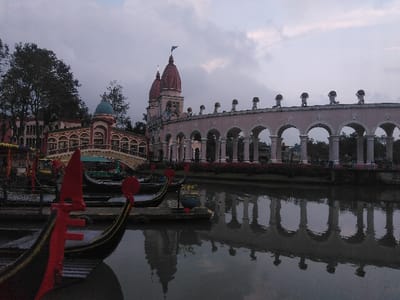 The image depicts an artificial canal scene in the Pueblo Antiguo area of Parque de Atracciones, Ciudad de San José, Costa Rica, under an overcast sky. In the foreground, a dark body of water reflects the surrounding architecture, showing ripples on its surface. Several gondola-style boats are moored at the left, with their pointed prows prominently visible; the closest boat features a red, upward-curving ornamental prow (ferro di prua) against a dark hull.
Along the canal's banks, ornate architectural structures are visible. To the left, a light-colored arched bridge with a teal-colored dome crosses over a section of the water, connecting to a multi-story building featuring multiple arches and classical design elements. This structure is partially obscured by dense, dark green tree foliage.
To the right, a lengthy, light-colored arcade structure extends horizontally along the water's edge. This arcade comprises numerous arches supported by pillars. Along its roofline, a series of evenly spaced statues or decorative figures, appearing to hold objects on their heads, are visible. Behind and to the right of this arcade, a large building complex features two prominent, reddish-brown onion-domed spires, with a dark pennant or flag atop the taller spire. The sky is uniformly gray and cloudy, indicating subdued light conditions. No people or overt actions are visible in the scene.