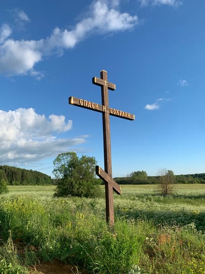 In the village of Svyatitsa, Russia, a wooden Orthodox cross stands in a field of wildflowers under a bright blue sky with scattered white clouds. The cross is dark brown with gold lettering on one of its beams. The lettering is in Cyrillic script and reads "СПАСИ И СОХРАНИ," which translates to "Save and Protect." The field is lush and green, with abundant white and yellow wildflowers. A dense forest can be seen in the background. The scene is peaceful and serene, evoking a sense of spirituality and natural beauty.