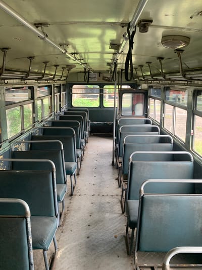 The image shows the interior of an old, empty bus in Koggala, Sri Lanka. The bus has rows of worn, teal-colored seats on both sides of a central aisle. The seats have metal frames and handles. Overhead, metal poles and straps for passengers to hold onto are visible. Sunlight filters in through the windows, illuminating dust particles in the air. The floor is a metallic diamond plate pattern. The environment outside the windows appears to be overgrown with greenery. The bus appears to be stationary and possibly abandoned or in storage. There are no people visible in the image.