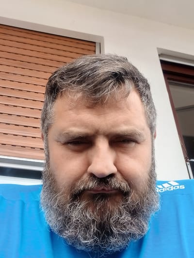The image is a close-up, head-and-shoulders portrait of a man with a full, graying beard and messy, graying hair. He is looking directly at the camera with his eyes partially closed. He is wearing a bright blue t-shirt with a white Adidas logo visible on the right side of his chest. The background consists of a brown slatted window blind on the left and a white wall with a dark-framed window on the right. The lighting suggests it is daytime. The image appears to be a selfie taken outdoors. There is no identifiable information in the image to pinpoint its location to Pfarrkirchen, Germany.