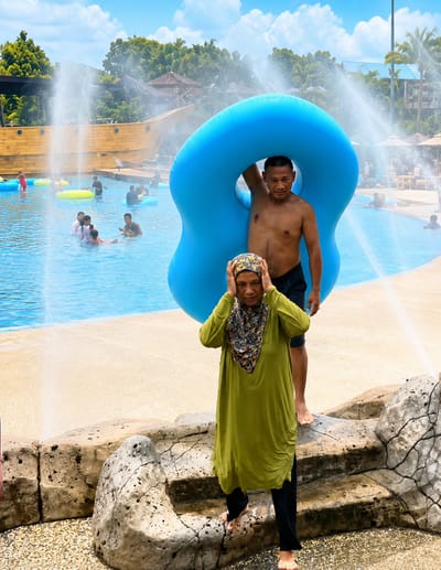 In the city of Eampat Kwalamencirim, Indonesia, at a water park, a man holding a large blue inflatable ring stands behind a woman wearing a green hijab and headscarf. The woman is looking down and holding her hands to her head. The man is shirtless and wearing dark shorts, with the blue ring obscuring his upper body. In the background, a pool is filled with people enjoying themselves, with a large wooden ship structure and water fountains adding to the lively atmosphere. The ground is wet, indicating the presence of water.