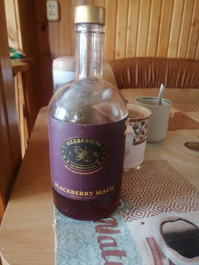 The image captures a close-up shot of a bottle of "HERBARISTA BLACKBERRY MAGIC" syrup, placed on a patterned tablecloth. The bottle has a metallic gold cap and a deep purple label with gold lettering. Next to the bottle, two white cups are visible; one is a decorative mug with a floral pattern, and the other is a plain white cup with a spoon inside. The background reveals wooden paneling, a cushioned seat, and what appears to be a wall outlet. The scene is set on a table covered with a tablecloth featuring a geometric design and a section of a paper placemat with a floral and text design. There are no people in the image, and the lighting suggests an indoor setting, possibly in a home or cafe. The overall impression is one of a still life arrangement.