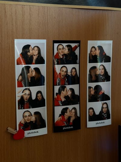 Three photobooth strips are on a wooden surface. The strips contain photos of two people, likely a couple, posing and kissing. The strips have "photobox." printed on them. One strip is secured with a small red heart-shaped clothespin clip. The location is not discernible from the image.