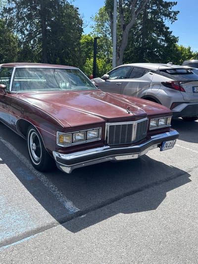 A dark red 1970s American sedan is parked in an asphalt lot under a clear blue sky. The car, with its distinctive chrome grille and rectangular headlights, is positioned in the foreground, partially obscuring a silver Toyota C-HR. Trees with green foliage are visible in the background, and the shadow of the sedan falls across the parking lot. The license plate of the sedan reads "74 ZJJ". The scene is likely a parking lot in Tabasalu alevik, Estonia, given the license plate format.