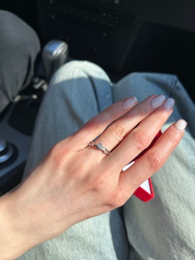 A close-up image depicts a human left hand adorned with a silver-colored ring featuring a single clear stone on the ring finger. The hand has a light skin tone, and the fingernails are manicured with a clear or light-colored base and subtle glitter tips. The hand rests on the upper thighs of an individual wearing light blue denim jeans. Partially visible beneath the ring finger and held by the thumb is a small, open red box, consistent with a jewelry box. In the blurred background to the upper left, a dark garment and a silver-colored automatic gear selector are visible, indicating the interior of a vehicle. The upper right background shows indistinct dark elements of the car's interior. The scene is illuminated by strong natural light from an upper-left source. The exact location within Călărași, Romania, cannot be determined from the image content.