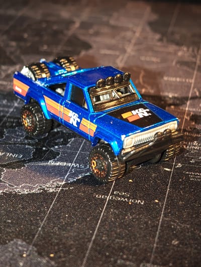 The image is a close-up, eye-level shot of a blue toy off-road pickup truck. The truck has orange stripes along the side and a "K&N" logo on the hood. It has large, knobby tires with bronze accents and off-road lights mounted on the roof. The truck is positioned on a dark, textured surface that appears to be a map, with faint lines and text visible. The lighting casts shadows on the map and highlights the metallic sheen of the truck. There are no people or animals present in the image. The location in the city of Dörpen, Germany, is not identifiable from the visual information provided.