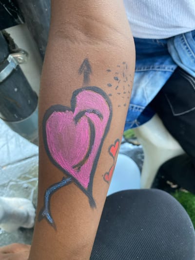 The image shows the lower right arm of an individual, likely a child, with a large pink heart painted on it. The heart is outlined in black and has a blue stalk extending from its base. Above the heart is a black arrow pointing upwards, and a cluster of small black dots. There are also two red hearts painted near the elbow. The individual is wearing a white t-shirt and blue denim jeans. The background is out of focus and partially obscured by a black fabric surface. The coordinates (latitude: 4.048, longitude: 9.218) correspond to a location in Bonadikombo, Cameroon. There are no other individuals in the image.