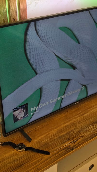 A television screen displays album art for the song "My Own Summer (Shove It)" by Deftones, featuring a coiled white snake on a green background. Below the song title, "Deftones" is visible. The television is placed on a wooden surface. To the left of the television, a black watch with a silver watch face and black strap lies on the same wooden surface. The lighting in the scene appears to be a mix of natural light and artificial light, with some sections of the background being illuminated by green and pink light sources. The location is identified as Liliak, Bulgaria.
