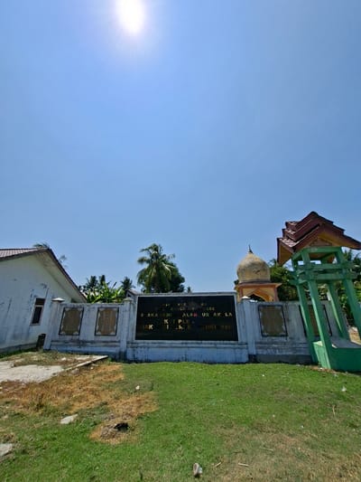 A wide-angle, outdoor, daytime, low-angle shot shows a monument or memorial in Kutakrueng, Indonesia. The monument is a white concrete wall with gold decorative panels on either side of a black plaque. The plaque has gold lettering and appears to be inscribed with text. Behind the wall, a palm tree and other foliage are visible. To the left of the wall is a white building with a red-tiled roof and a single window. To the right of the wall is a green, open-air structure with a red-tiled roof. In the background, a dome-shaped structure is visible, as are more palm trees. The sky is a bright, clear blue, with the sun prominently displayed in the upper left corner, causing lens flare. The ground in front of the monument is covered in green grass, with some patches of dry, brown earth.