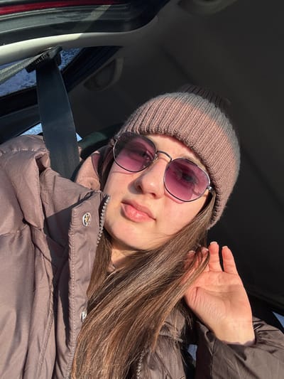 A young woman wearing sunglasses and a knit hat poses inside a car. She's wearing a brown puffer jacket and a seatbelt. Her hand is raised near her head. The lighting is bright, creating strong shadows on her face and clothing. Her long brown hair is shiny and reflects the light. The car's interior is visible, including the door frame and a glimpse of the outside.