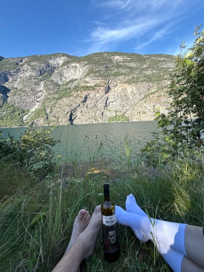 An outdoor, eye-level shot captures a serene moment by a body of water, likely the Lærdalsfjorden near Lærdalsøyri, Norway. Two individuals' bare feet and lower legs are visible in the foreground, resting on a grassy embankment. One person's feet are bare and show sparse leg hair, while the other person's legs are clad in white socks. A bottle of "19 Crimes The Uprising" red wine stands upright in the grass between the individuals. The background features a calm, greenish-blue body of water, leading to a steep, rocky mountainside covered in scattered trees. The sky above is a clear, bright blue with wispy white clouds. The scene suggests a leisurely outdoor activity in a natural setting.