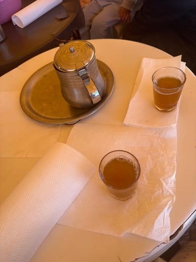 Two glasses of liquid, likely tea, sit on a round, light-colored table, each atop a square of crinkled white paper. A metal teapot with a lid and a metal tray underneath are positioned on the left side of the table. A roll of paper towels lies partially on the table’s surface. In the background, blurred figures suggest the presence of people, one possibly wearing a light-colored garment. The scene is in Boufakrane, Morocco, as it includes Moroccan tea.