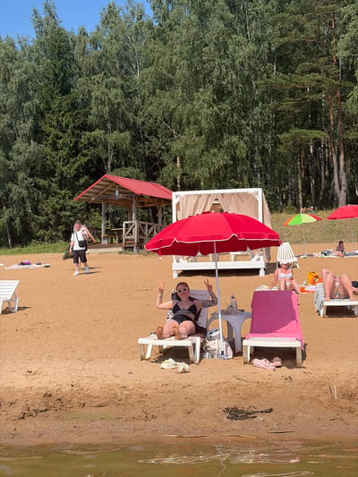 The image depicts a sunny day at a sandy beach, likely in Ozyaritska-Slabadski rural council, Belarus, given the location context. The scene is a summer leisure setting with several people relaxing on beach chairs. A prominent red umbrella shades a section of the sand, and a smaller, colorful umbrella is also visible. Two white canopies, possibly part of a beach club or relaxation area, are set up on the sand. In the background, a dense forest of green trees borders the beach, with a clear blue sky above.
Several individuals are captured in various states of relaxation. One person is lying on a beach chair in a black bikini, striking a peace sign with their fingers and wearing sunglasses. Another person in a white bikini is sitting on a beach chair, seemingly posing. Further back, other beachgoers can be seen lounging on chairs, some partially obscured by umbrellas or other beach furniture. A young man in a white t-shirt and black shorts is standing on the sand, looking towards the camera. The overall mood appears to be relaxed and enjoyable, consistent with a summer vacation or day at the beach. The lighting suggests it is midday, with bright sunlight casting shadows on the sand. The water of a lake or river is visible on the right side of the image, reflecting the sunlight and surrounding greenery. There is no visible text in the image.