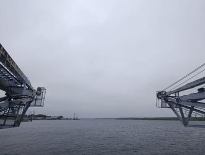 The image displays a wide, grey-blue body of water under a uniformly overcast, light grey sky in Igrim, Russia. In the immediate foreground, large, weathered industrial structures frame the view, extending diagonally from the upper left and upper right corners towards the center. The left structure is a heavy metal framework of girders and grates, showing signs of rust, indicative of a conveyor or gantry system. A similar, symmetrical metal structure occupies the right foreground, also composed of heavy girders, grates, and featuring a large pulley mechanism at its visible end, suggesting another part of a large industrial loading or unloading apparatus.

Across the water on the left shore, a low-lying industrial complex is visible, comprising several buildings, some with distinctive blue roofs, and a cluster of multiple cranes, including tall gantry cranes and mobile cranes, indicating port or cargo handling activity. Sparse green vegetation surrounds these structures. The distant right shore is marked by a continuous, dense line of dark green trees or vegetation. The water itself is dark blue-grey with subtle ripples, reflecting the muted light from the sky. No people or animals are visible in the scene. The overall lighting is diffused and flat due to the heavy overcast conditions.