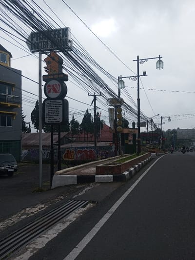 A street view features a tangle of overhead wires dominating the sky. A tall sign with directional information and cartoon graphics stands to the left. The road is asphalt with white line markings, and a raised median with vegetation runs along the street. The sky is overcast, and a few motorbikes are visible further down the road. Graffiti adorns a wall in the background, partially obscured by trees.