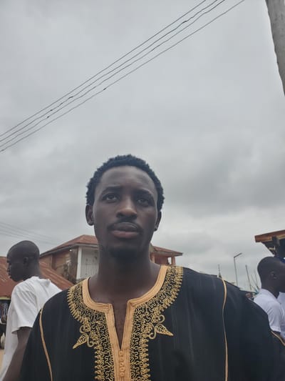A medium shot captures a Black man with short, curly hair and a mustache. He is wearing a black garment with intricate gold embroidery around the neckline and down the front. The man is looking upwards and to his right. In the background, another Black man in a white t-shirt is visible, walking away from the camera. Behind him, buildings with corrugated metal roofs suggest an urban or semi-urban environment. Overhead, power lines stretch across a cloudy sky. The overall scene appears to be an outdoor setting in Ohit, Nigeria.