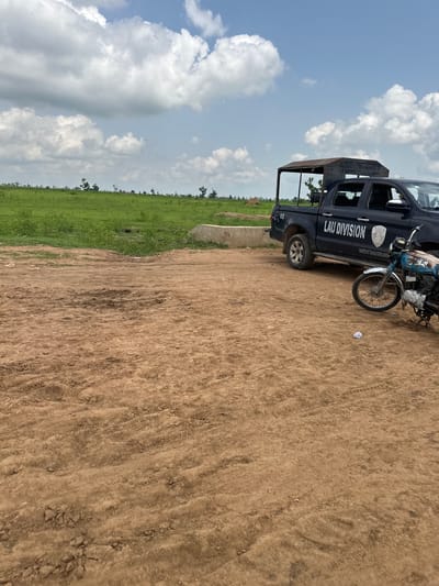 The image depicts an outdoor scene in Garin Dogo, Nigeria. A black pickup truck, identified with the markings "LAU DIVISION" and "02", is parked on a dirt surface. A blue and white motorcycle is positioned to the right of the truck. The background features a vast expanse of green grass under a sky with scattered clouds. The terrain appears to be undeveloped or rural.