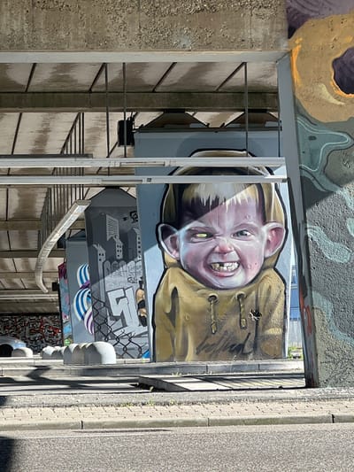 The image captures an urban underpass or concrete structure in Weert, Netherlands, extensively covered in vibrant street art under bright daylight.
The dominant feature is a large, detailed graffiti mural of a person's face, likely a child or young adult, with light-colored hair and prominent ears. The face displays a wide, mischievous grin, revealing teeth, and large, expressive eyes. The person appears to be wearing a brown or khaki-colored jacket. The style is a blend of realistic rendering with exaggerated, cartoonish features, giving the character a playful yet intense expression.
Below this central mural, another graffiti piece depicts a grayscale cityscape with stylized buildings, partially obscured by fencing patterns. Overlaid on these buildings are stylized letters, appearing as tags, with "GTS" being partially discernible. Further down, the wall features wavy blue and purple patterns, alongside more abstract and colorful graffiti designs.
The scene is framed by rough, textured concrete walls and pillars, typical of an underpass environment. Various metal railings and bars are integrated into the structure, with a distinctive curved metal rail extending into the lower-middle foreground. Bright sunlight casts sharp shadows, indicating clear weather during the day. No specific activity or event is taking place, as the image focuses on the static display of public art.