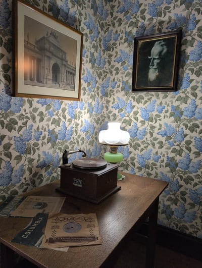 The image depicts an indoor scene with floral wallpaper featuring blue blossoms and green leaves. On the left, a framed black and white photograph of an arched building with columns is mounted on the wall. To the right of the photograph, a framed portrait painting of a man's profile, rendered in shades of blue and gray against a dark background, hangs on the wall. In the foreground, a wooden table holds a vintage record player, a table lamp with a white glass shade and a green base, and several records. One record is in a brown paper sleeve with text that reads "RCA Victor Company, Inc. Camden, N.J. U.S.A." Another record sleeve is visible with the word "COLUMBIA" printed on it. The lighting appears to be ambient, with a slight glow emanating from the lamp. The overall impression is of a preserved or historically styled room. There are no people visible in the image.