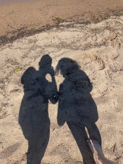 This image depicts a sandy beach scene, likely a shoreline, in Belozersk, Russia. Two distinct shadows are cast on the light brown sand, indicating the presence of two individuals. The shadow on the left appears to be holding a device, possibly a camera, suggesting they are taking a photograph. The shadow on the right is slightly closer to the viewer and shows the outline of a person standing. In the lower right corner of the image, a bare foot with painted toenails is visible, also casting a shadow. The beach is littered with small debris such as twigs, leaves, and pebbles. The water's edge is visible in the upper portion of the image, indicated by a darker, wet sand line.